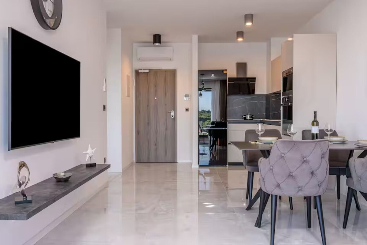 A modern kitchen with dark cabinets, a dining area with a table and chairs, and a flat-screen TV mounted on the wall.