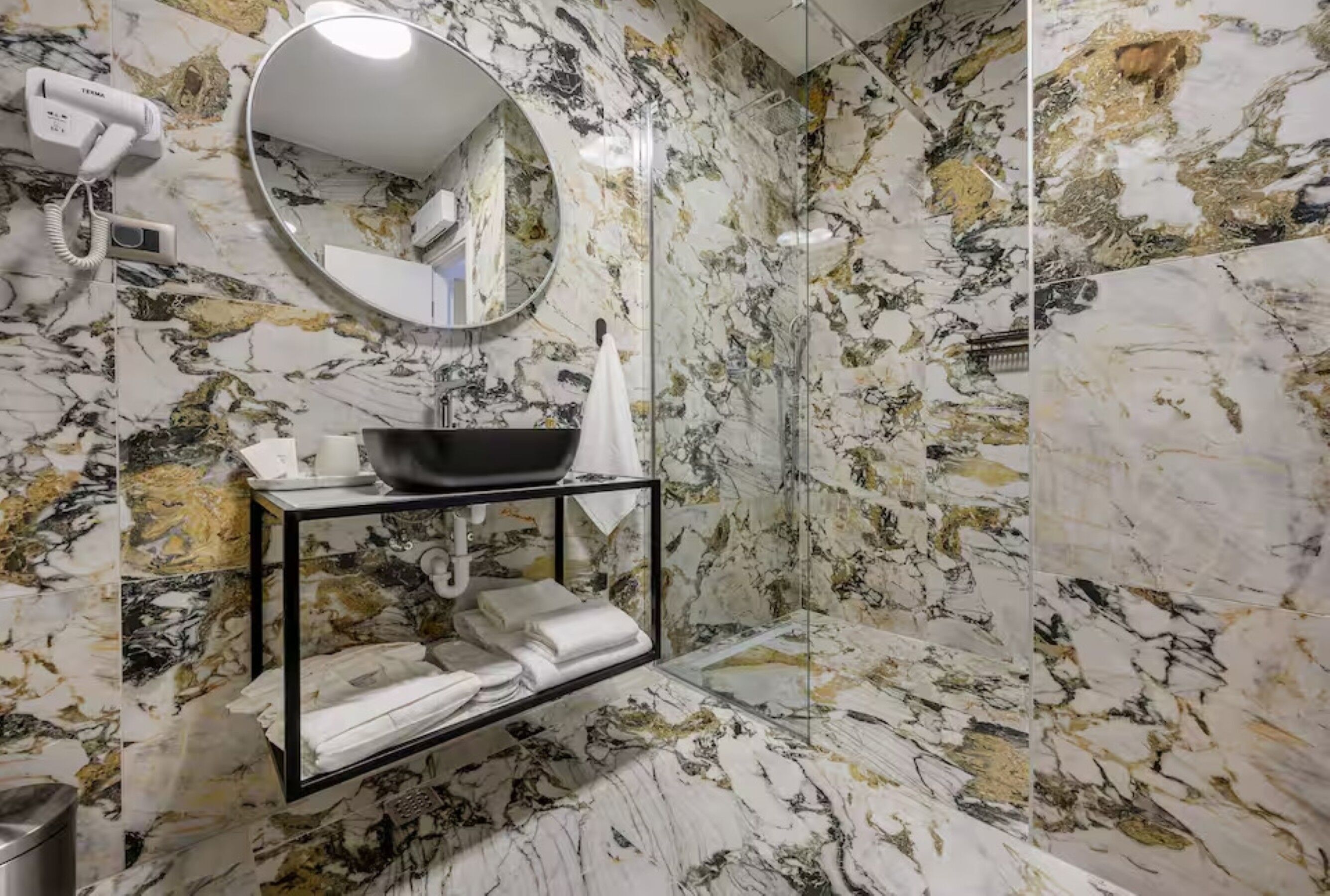 Une salle de bain avec un mur en marbre, un miroir rond, un lavabo avec un receveur noir et un porte-serviettes.