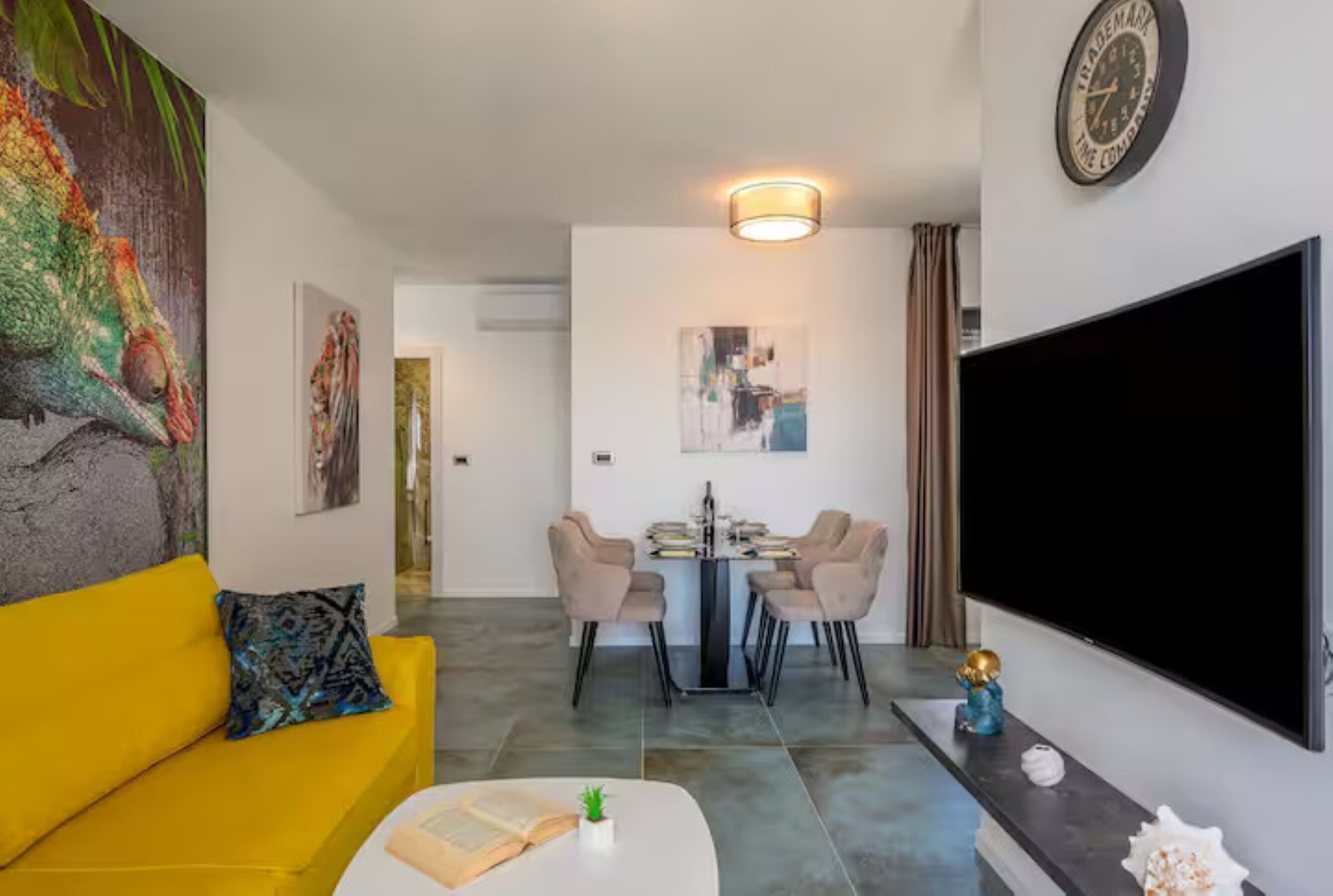 A living room with a yellow sofa, a dining area with a table and chairs, a flat-screen TV, and a large painting on the wall.