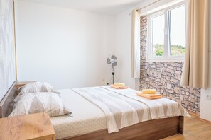A bedroom with a bed, a bedside table, a fan, a window with curtains, and a stone wall. - Apartment Adriatic Panorama (Podstrana)