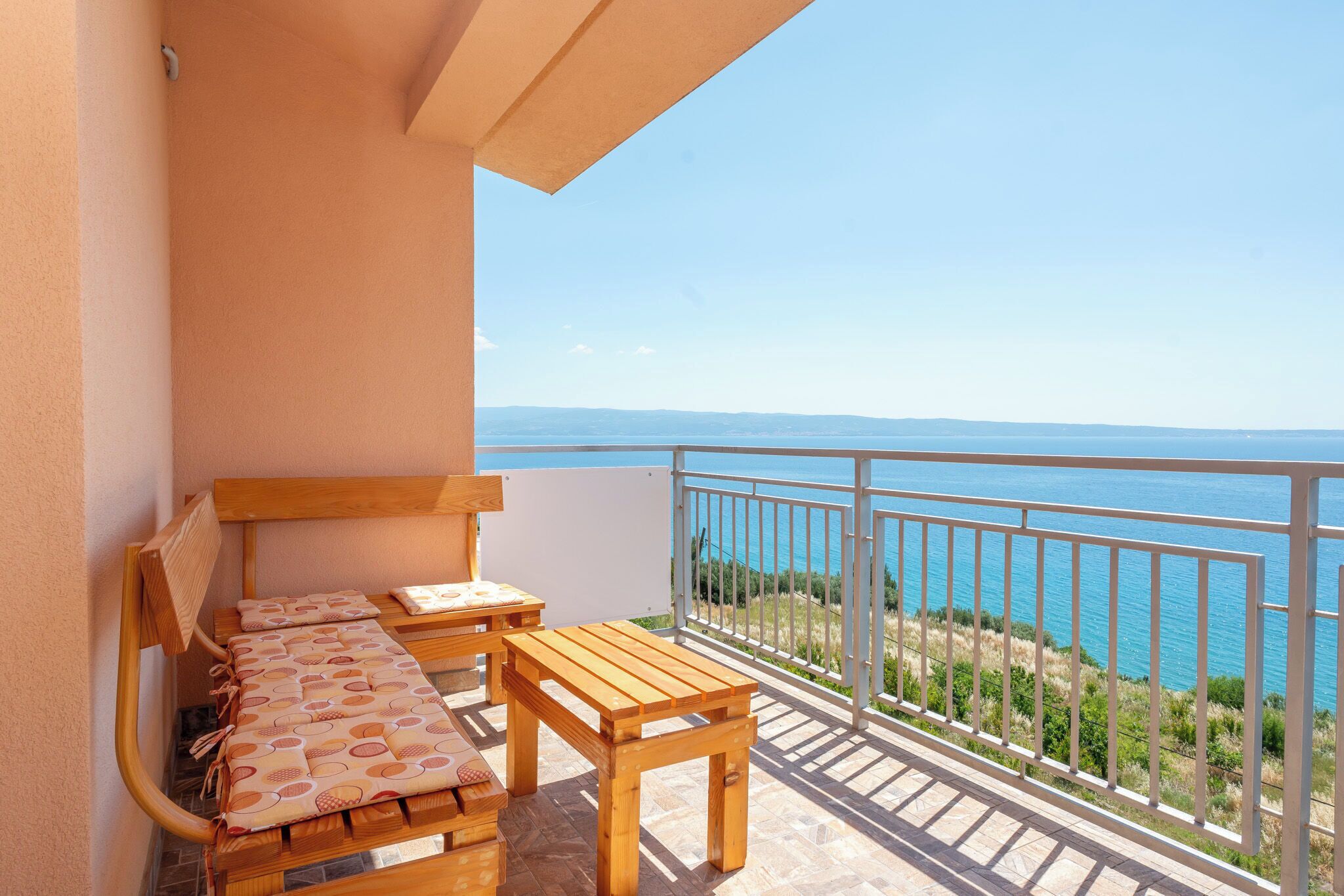 Een balkon met een houten tafel en bankje, een witte muur en uitzicht op zee.