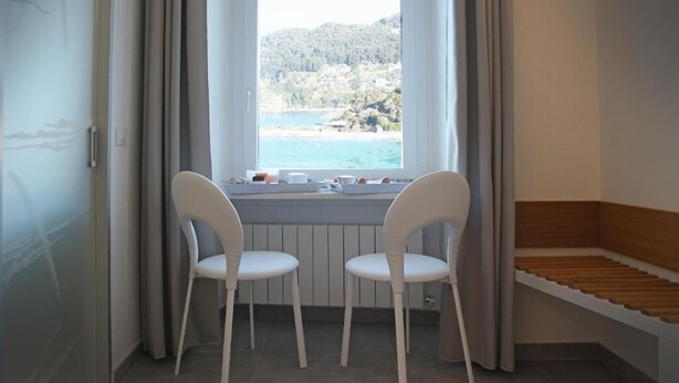 A room with a window overlooking a beach, two white chairs, a wooden bench, and a radiator.