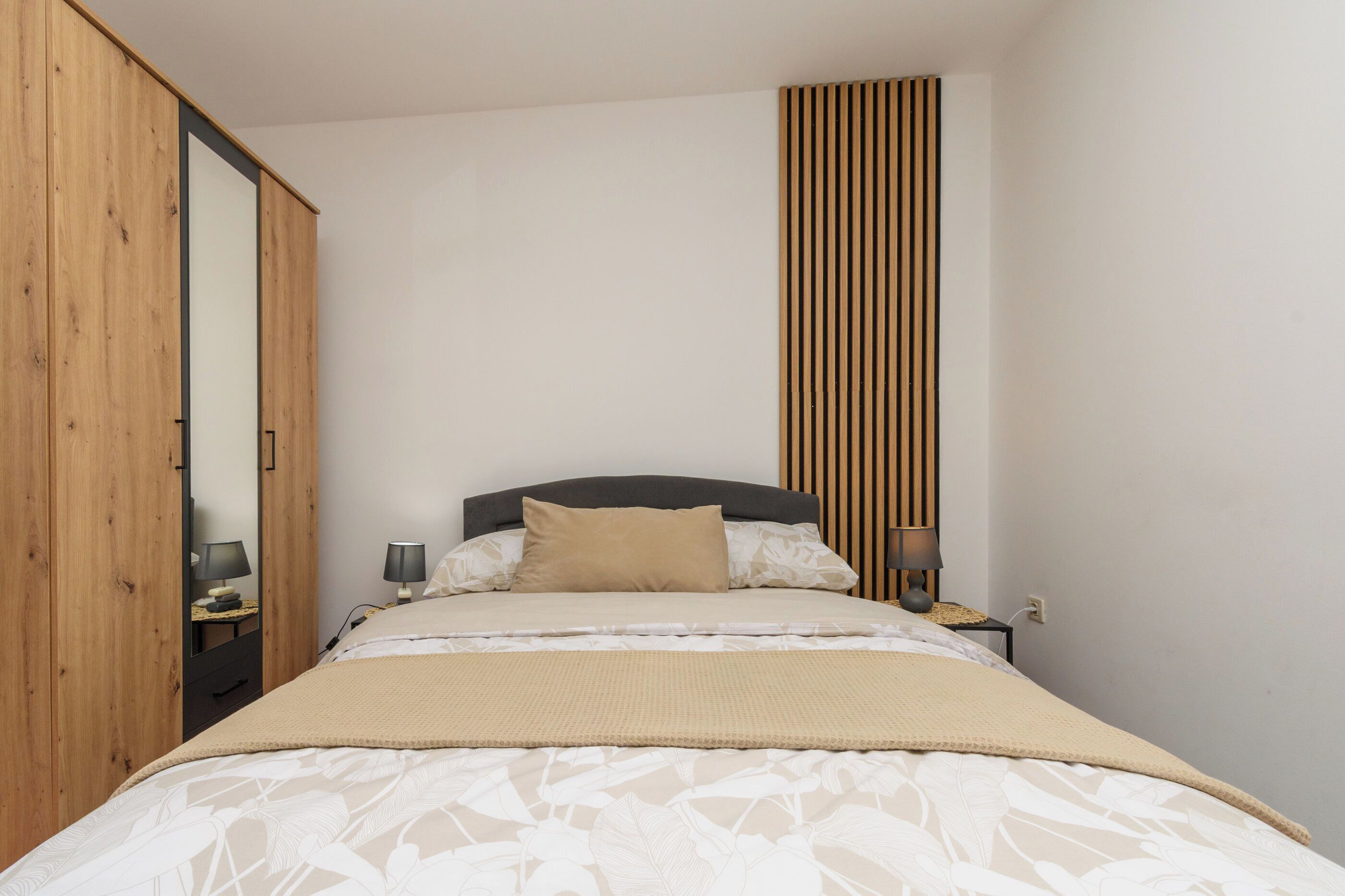A bedroom with a bed, a nightstand, a lamp, a mirror, and a wardrobe.