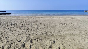 Ein sandiger Strand mit Fußabdrücken, die zum Wasser führen, ein Steg links und ein klarer, blauer Himmel.