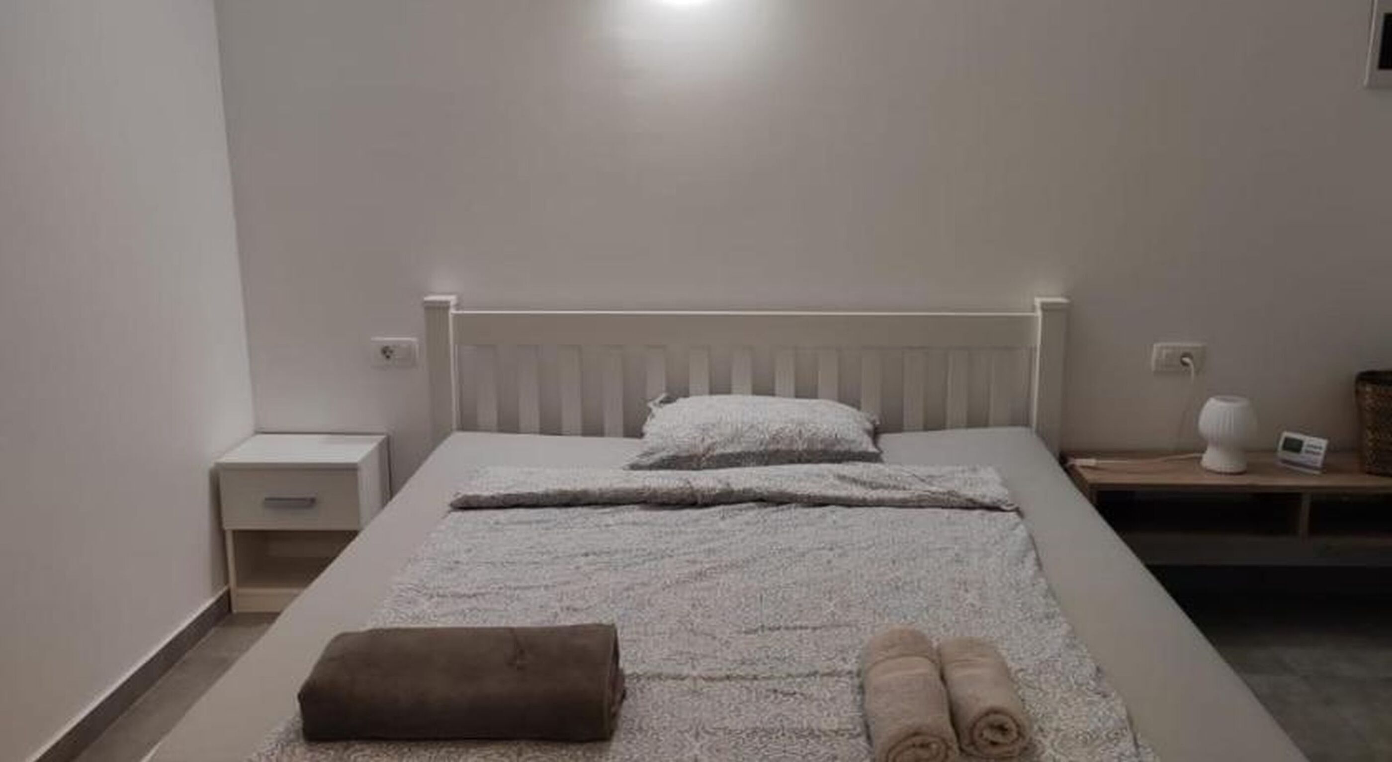 A neatly made bed with a white headboard, a bedside table with a lamp, and a small nightstand with a clock.