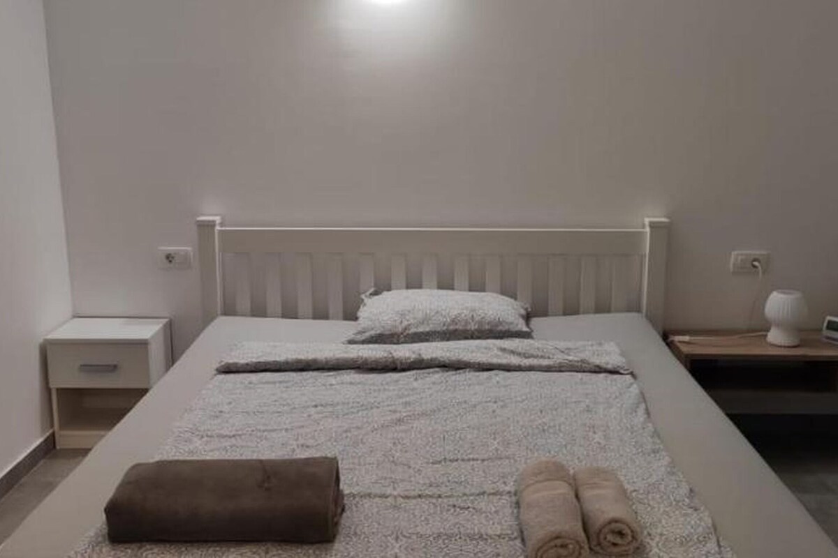A neatly made bed with a white headboard, a bedside table with a lamp, and a small nightstand with a clock.