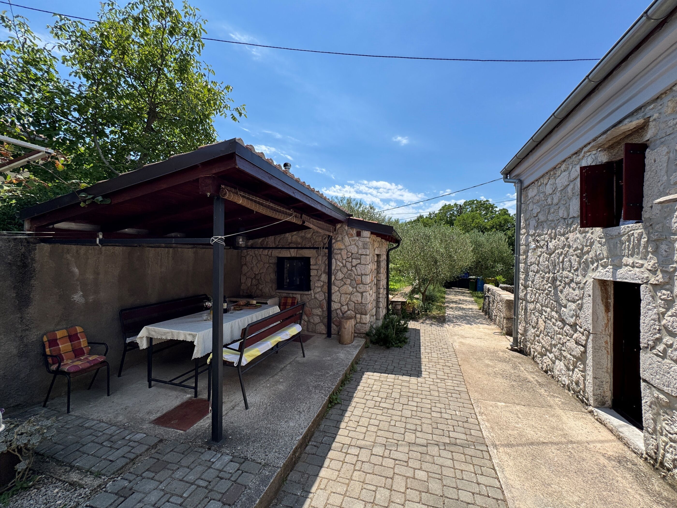 Un edificio de piedra con un patio cubierto, una mesa con sillas y un sendero estrecho que lleva a otro edificio.