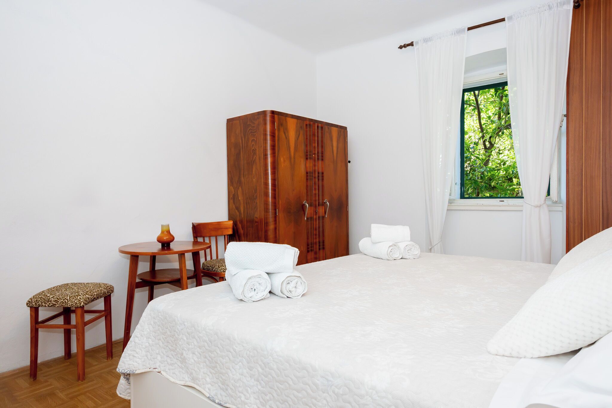 A bedroom with a bed, a wooden wardrobe, a small table with a vase, and a chair.