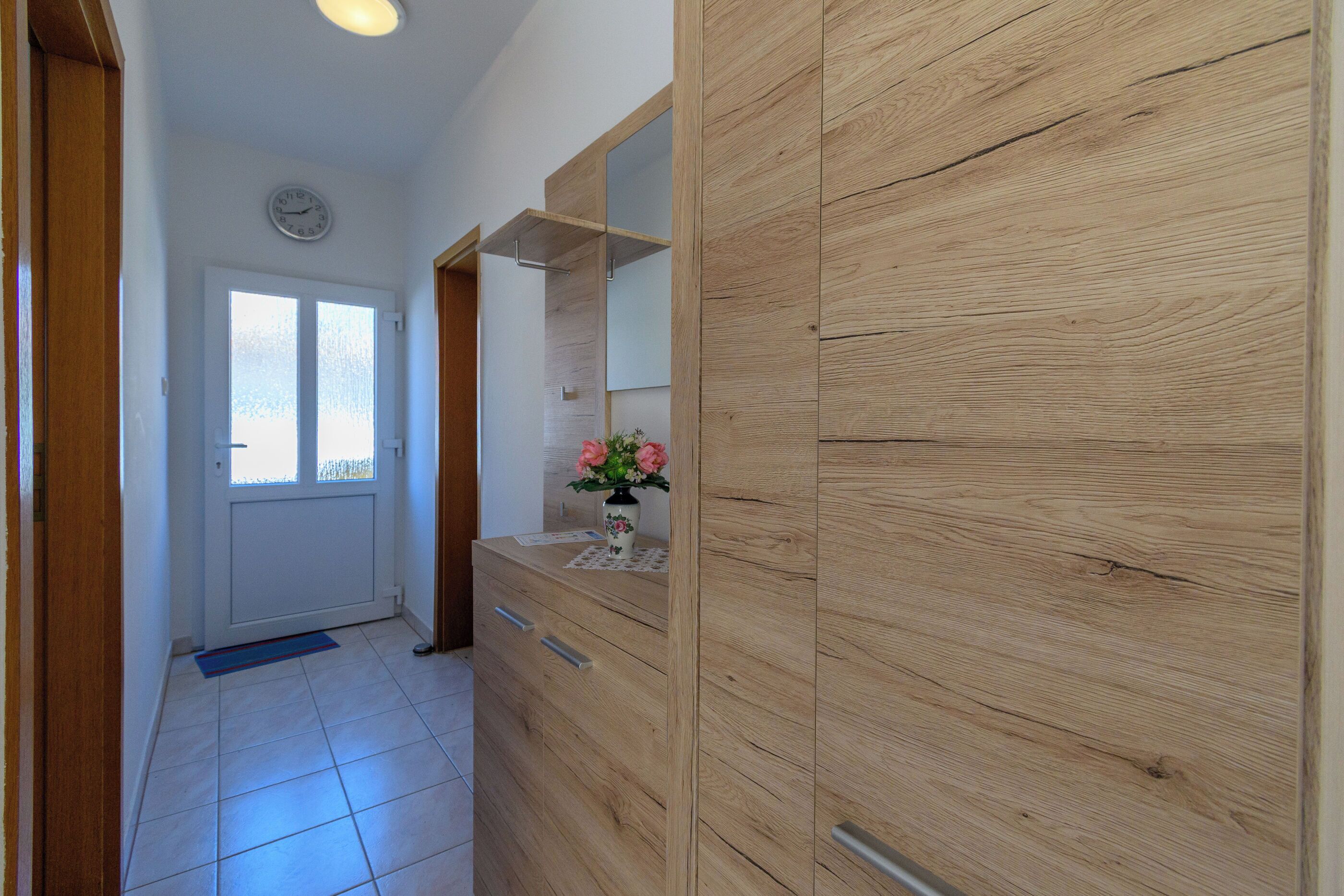 Un corridoio con un'armadiatura in legno, uno specchio, una sveglia appesa al muro e una porta con una finestra.