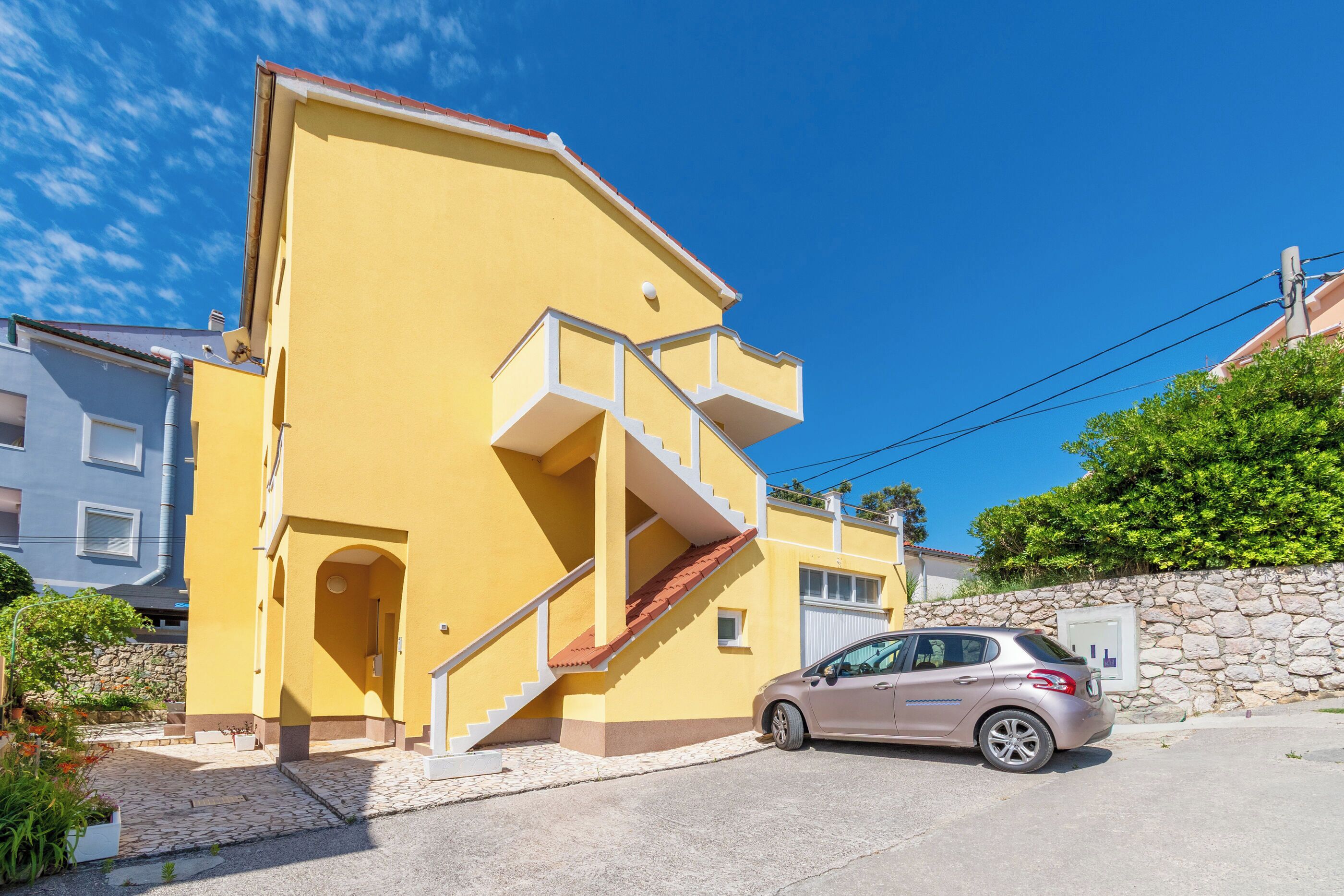 A yellow building with a car parked in front, a staircase, and a stone wall.