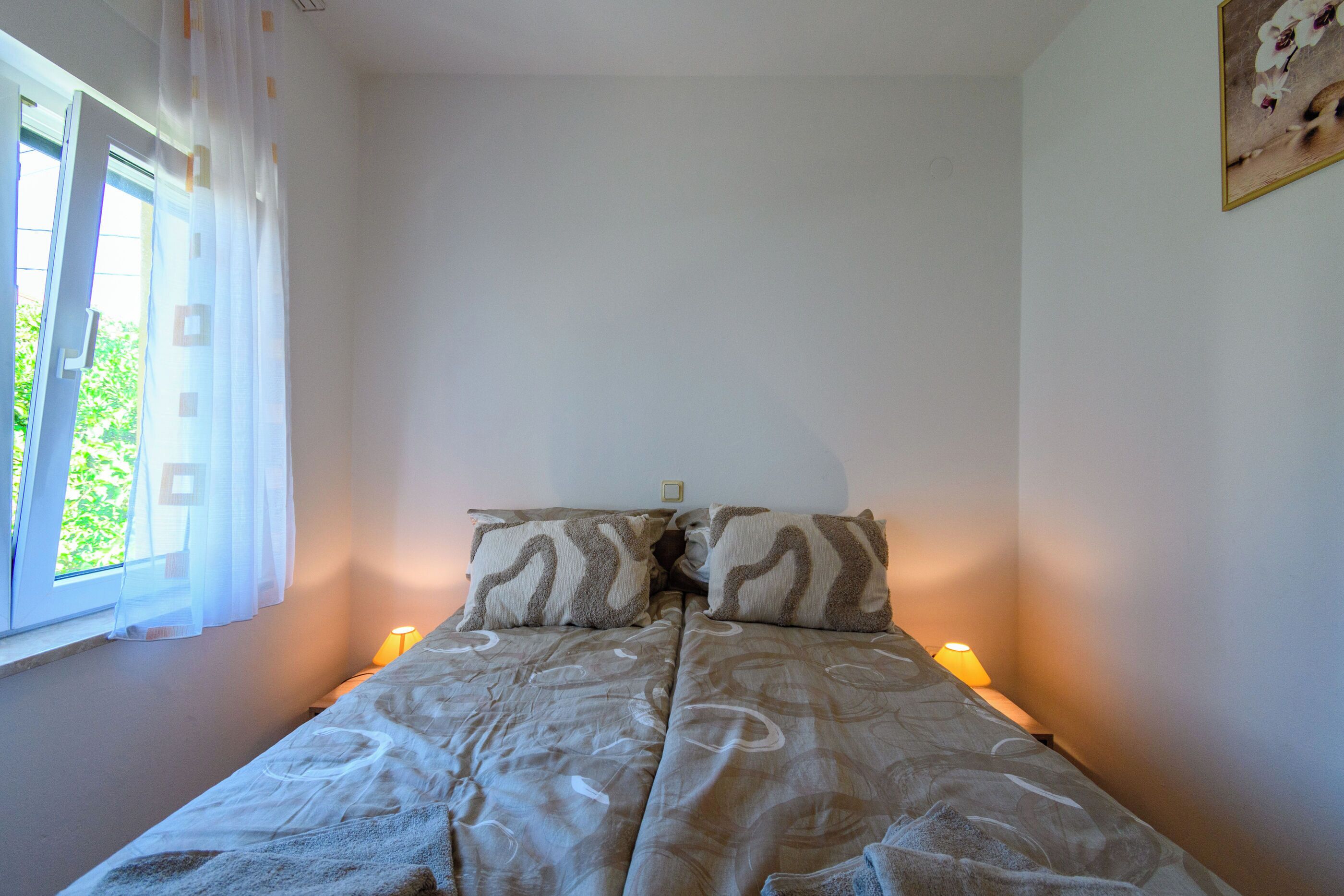 Une chambre à coucher avec un lit, deux oreillers, une table de chevet avec une lampe, une fenêtre avec des rideaux et un tableau accroché au mur.