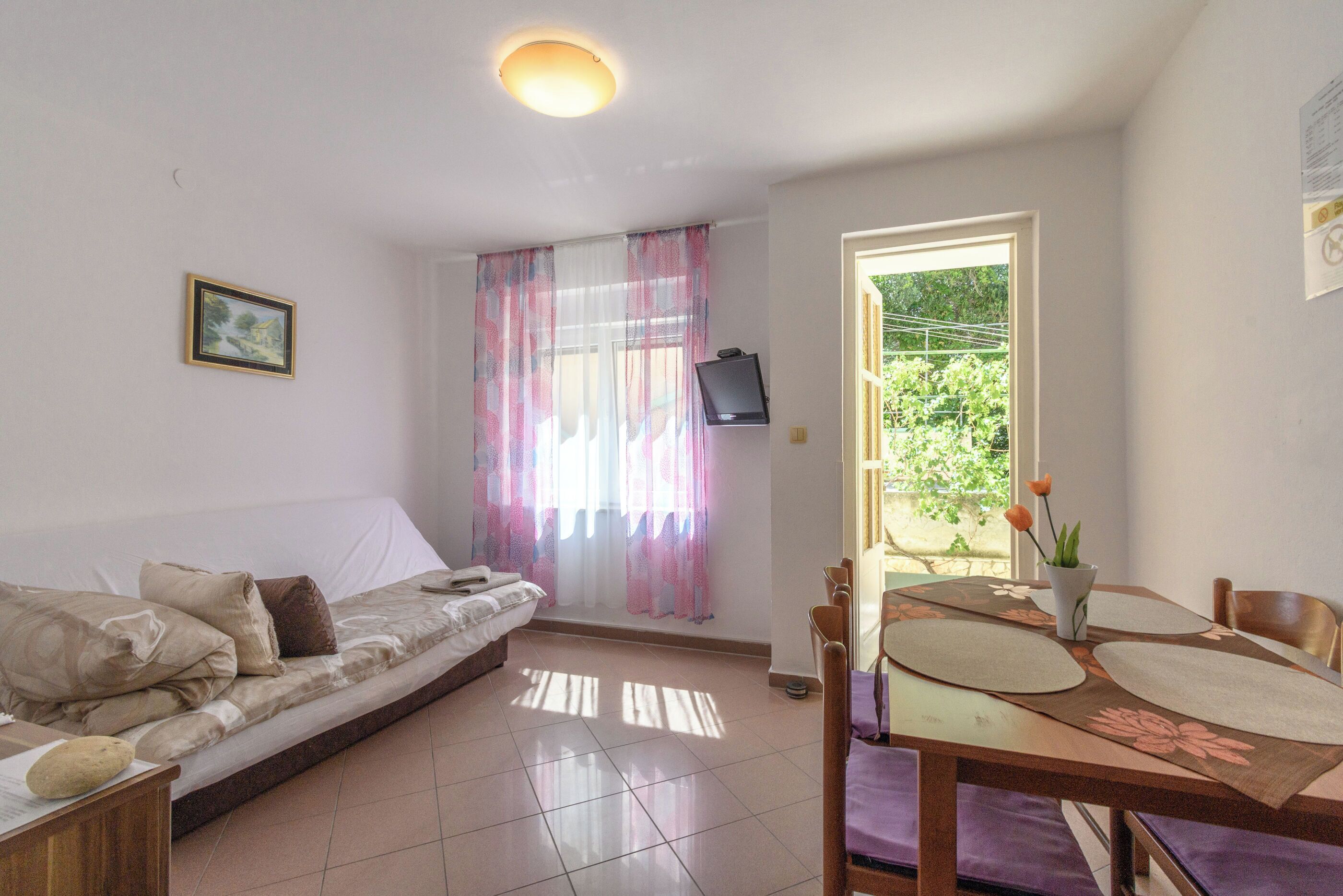 Una habitación con sofá, una mesa de comedor con individuales, televisor en la pared y ventana con cortinas.