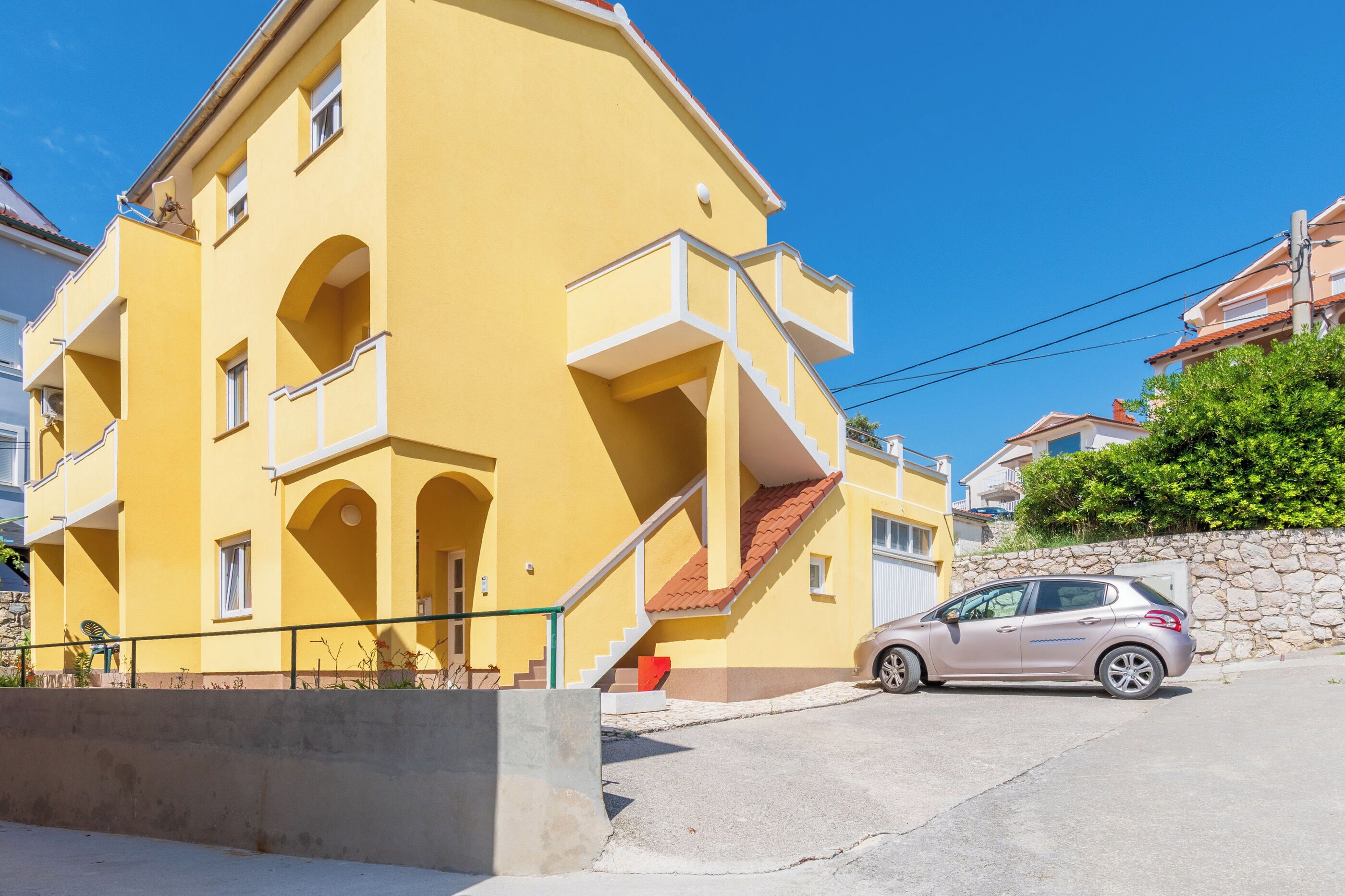 A yellow multi-story building with balconies, a car parked in front, and a stone wall to the right.