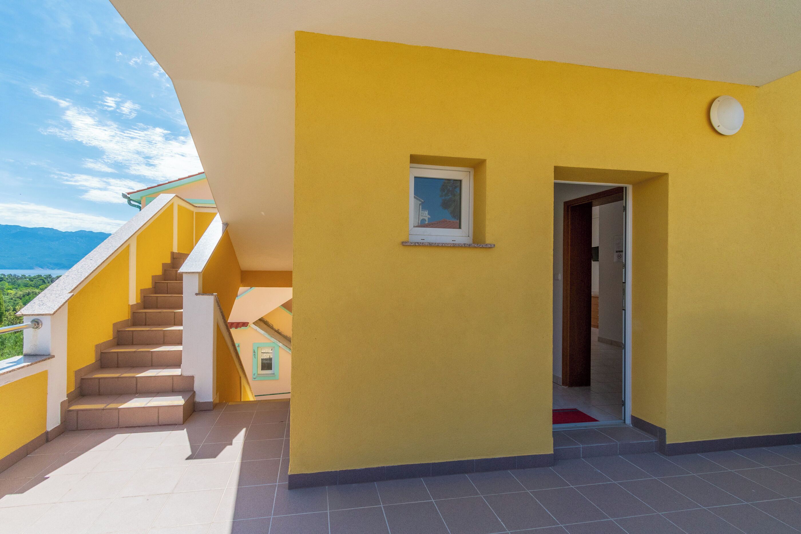 Un bâtiment jaune avec un escalier menant à un balcon, une petite fenêtre et une porte.
