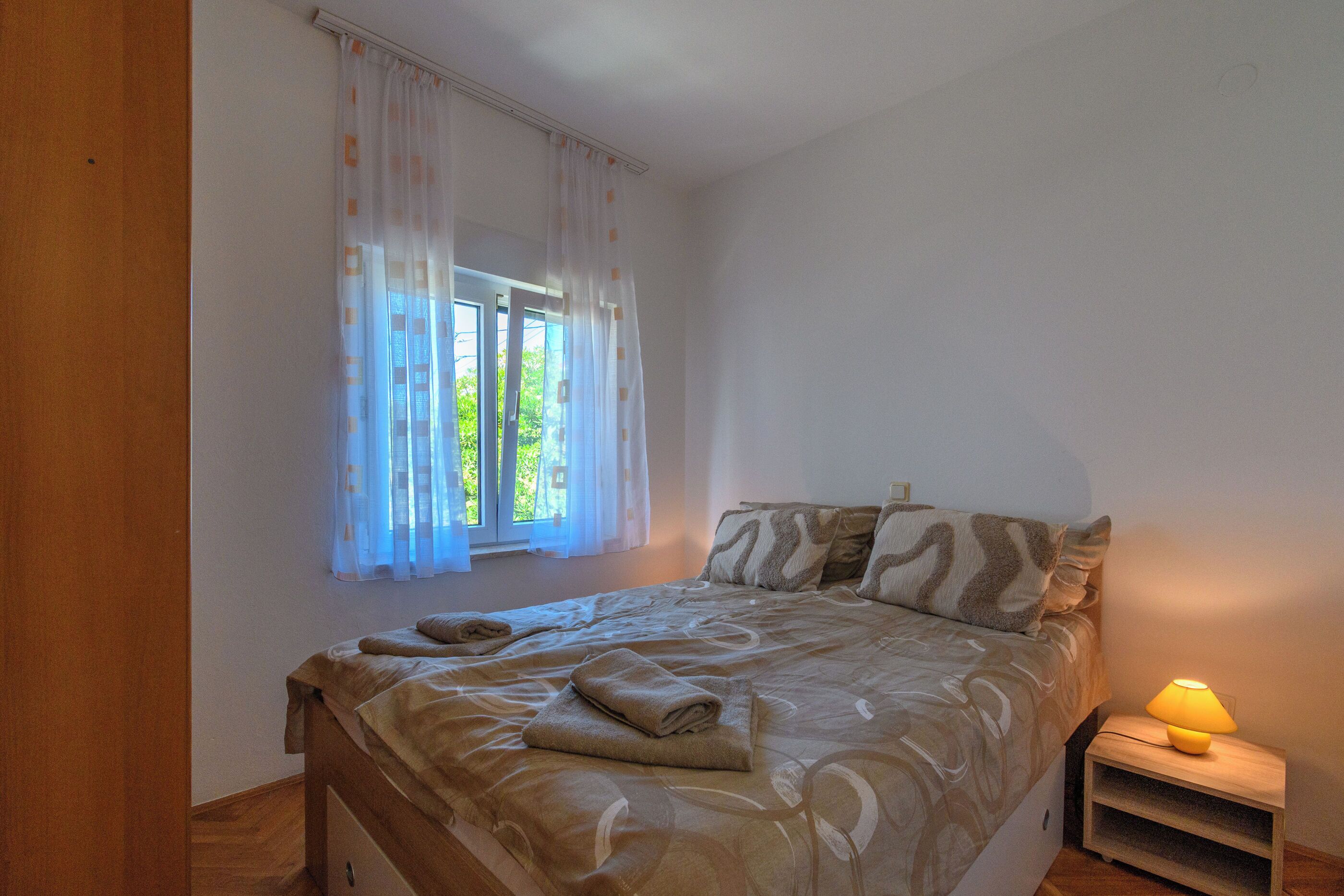 A bedroom with a bed, a bedside table with a lamp, a window with curtains, and a wooden floor.