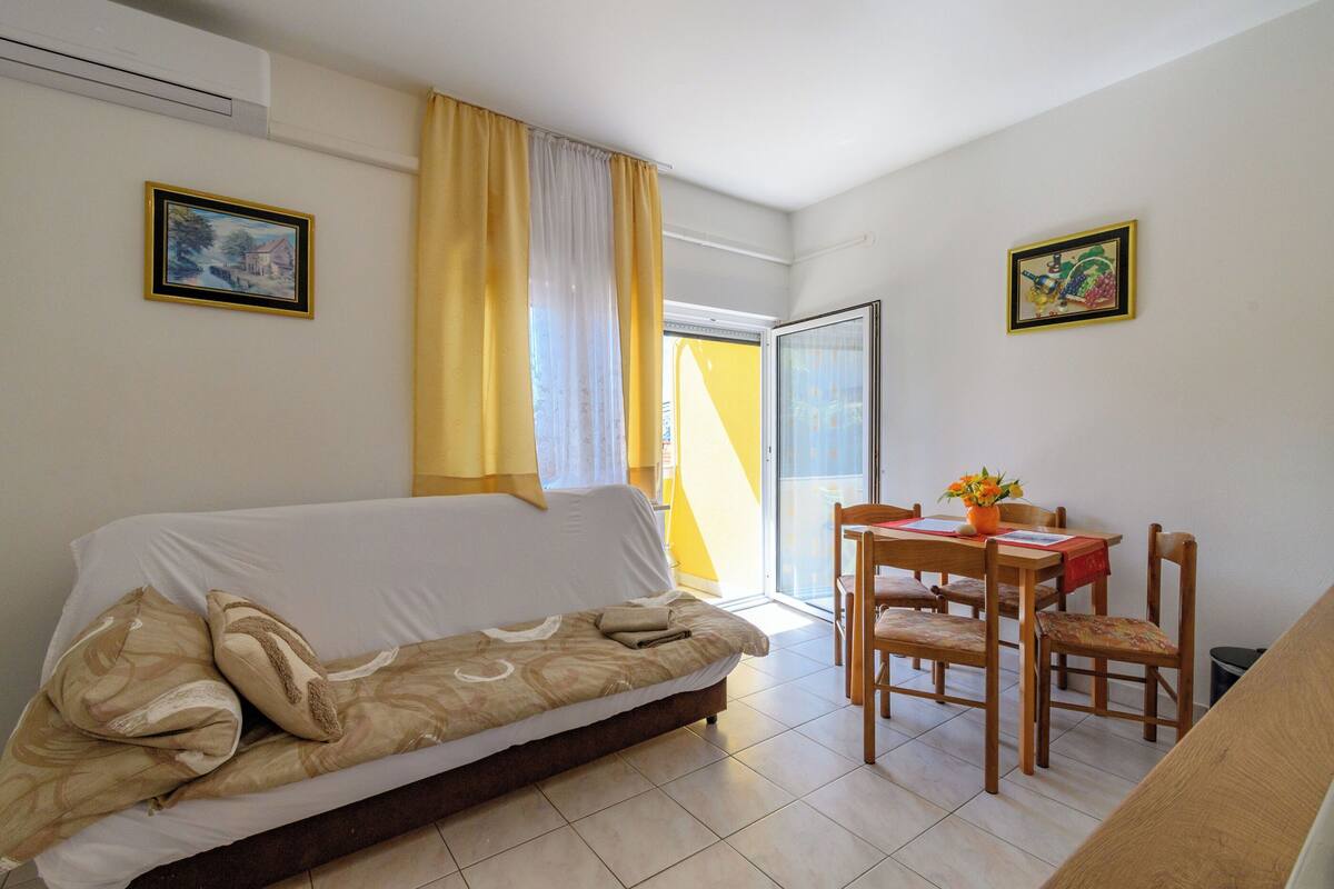 Ein Zimmer mit einer Couch, einem Esstisch mit Stühlen, einem Fenster mit gelben Vorhängen und zwei gerahmten Bildern an den Wänden.