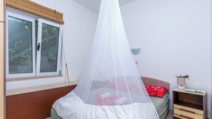 A bedroom with a bed under a mosquito net, a bedside table with a lamp, and a window with a view of trees. - Apartments Zeljana on the Beach (Podgora)