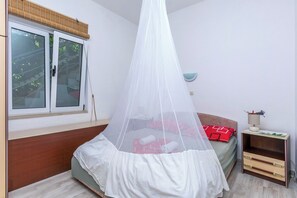 Ein Schlafzimmer mit einem Bett, das von einem Moskitonetz verhüllt ist, einem Nachttisch mit Lampe und einem Fenster mit Blick auf Bäume.