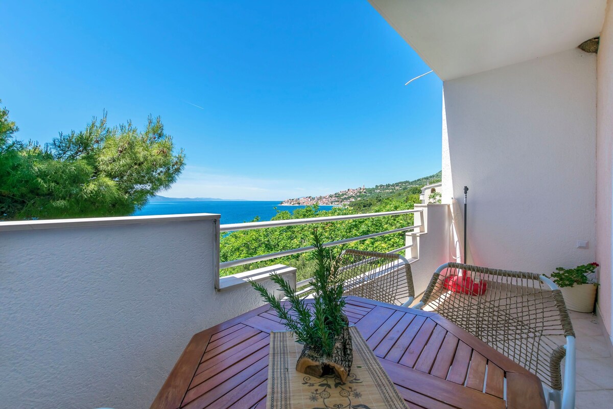Ein Balkon mit Holztisch, Topfpflanze und Blick aufs Meer.