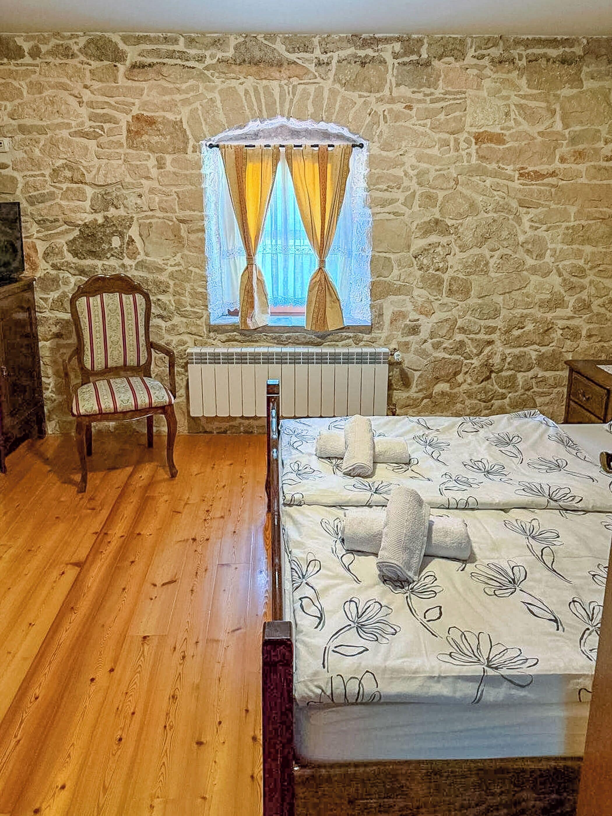 Ein Schlafzimmer mit Steinsturz, Holzstuhl, Heizkörper, einem Bett mit floralem Bettbezug und einem Fenster mit Vorhängen.