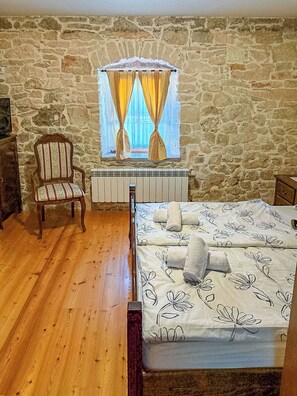 Ein Schlafzimmer mit Steinsturz, Holzstuhl, Heizkörper, einem Bett mit floralem Bettbezug und einem Fenster mit Vorhängen.