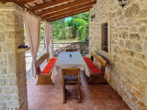Een overdekt terras met stenen muren, een tafel, banken en uitzicht op groen.
