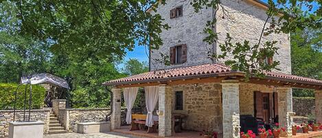 A stone building with a tiled roof, a covered patio with outdoor seating, and a garden with potted plants.