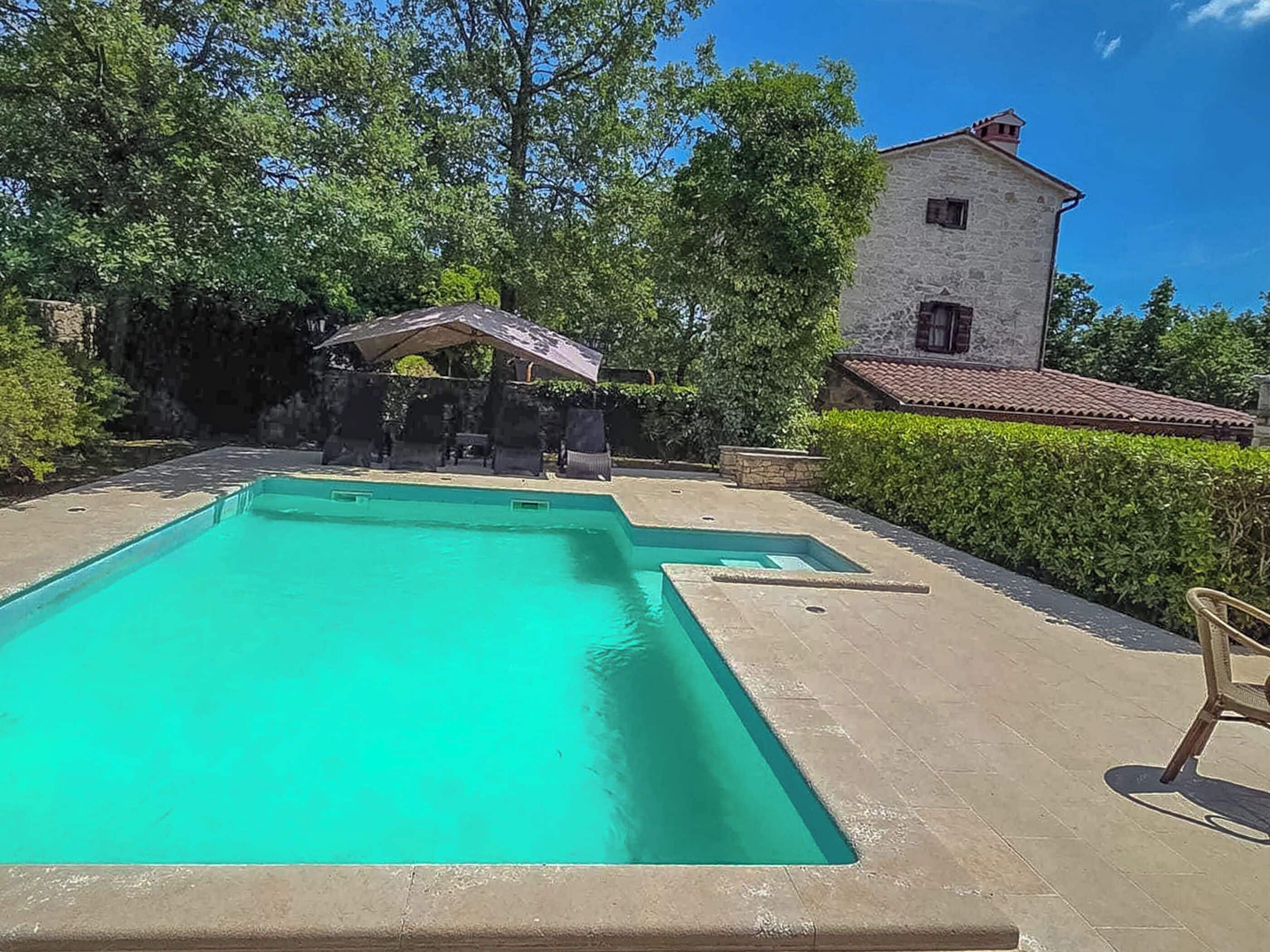 Ein rechteckiger Pool mit einer Steindeck, umgeben von Grünflächen und einem Steinhaus mit einem Ziegeldach.