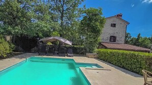 A rectangular swimming pool with a stone deck, surrounded by greenery and a stone building with a tiled roof. - House Stancija Bursic (Marcana)