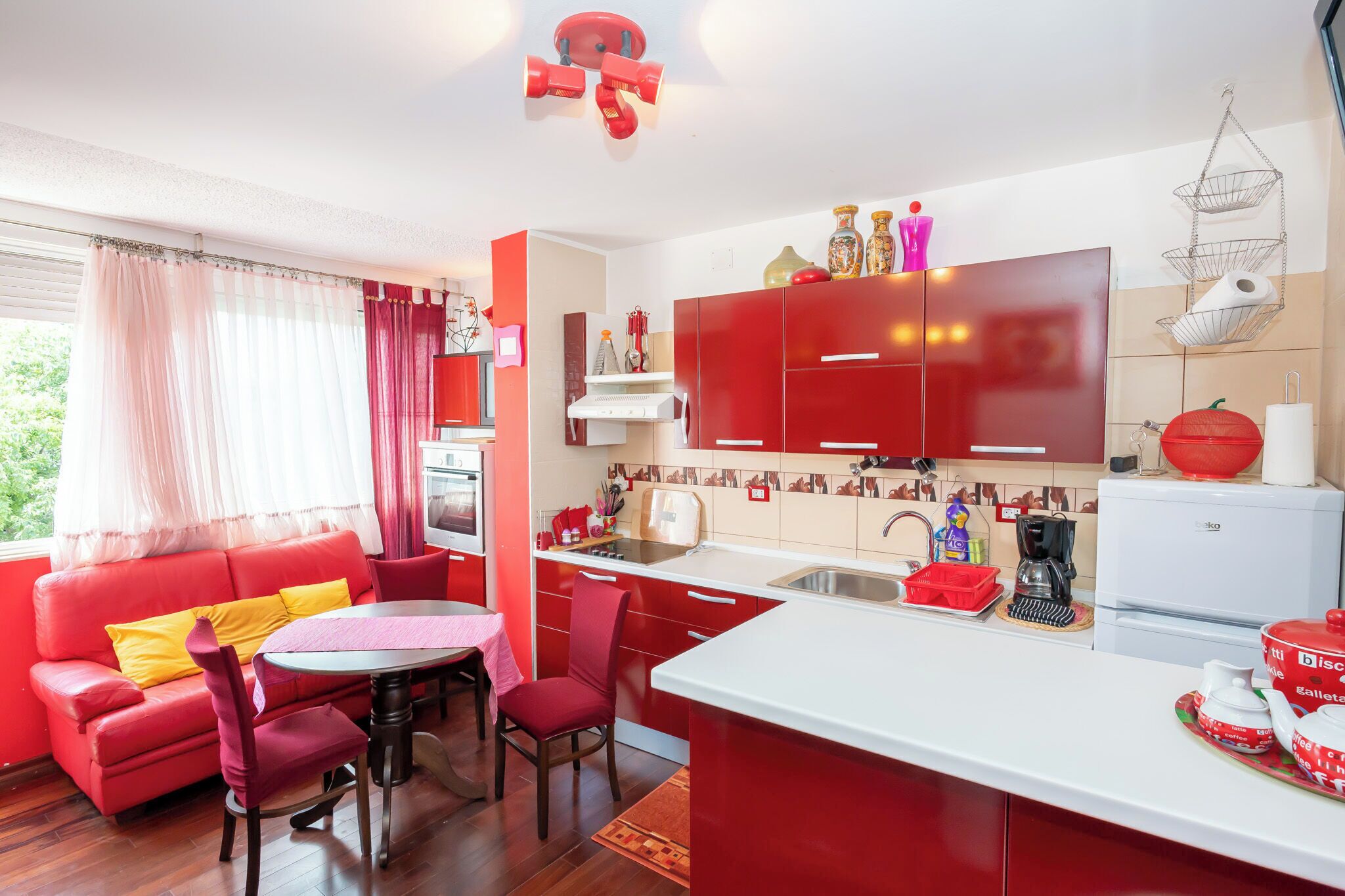 A compact kitchen with red cabinets, a dining area with a red sofa and chairs, and a small island with a sink.