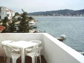 A balcony with a white table and chairs, overlooking a body of water with boats and buildings in the background. - Apartments and Room Gulic (Rab)
