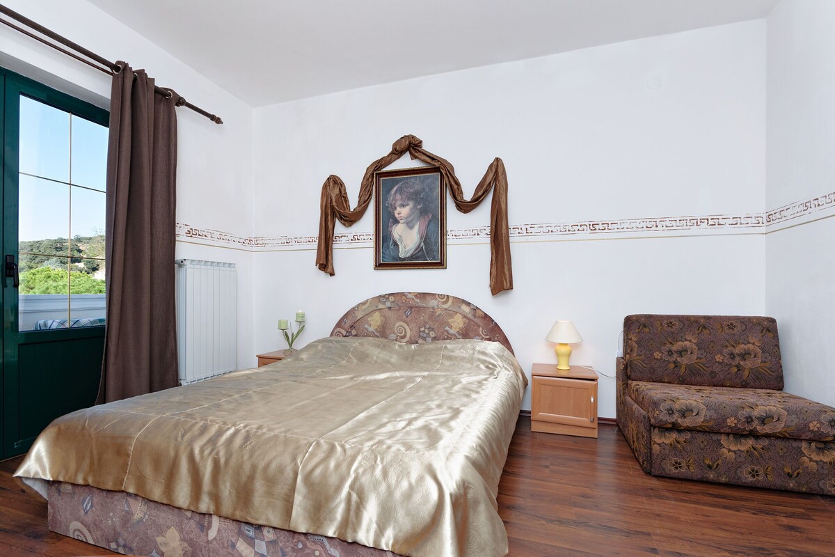A bedroom with a bed, a chair, a nightstand, a lamp, a painting, and a window with curtains.
