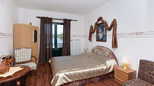 Quarto de hotel com cama, uma cadeira, guarda-roupa de madeira, janela com cortinas, radiador, quadro emoldurado e abajur numa mesa de cabeceira.