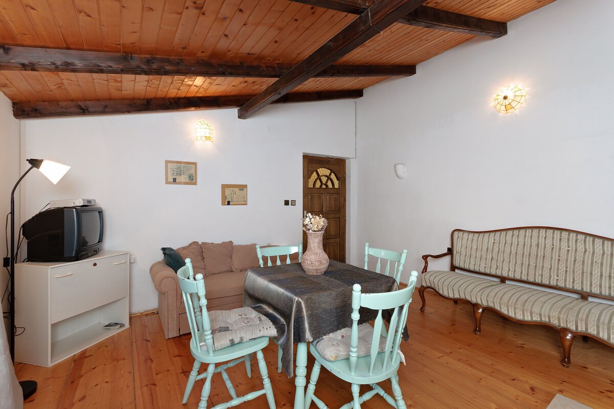 A cozy room with a wooden ceiling, a television on a cabinet, a sofa, a dining table with chairs, and wall-mounted lights.