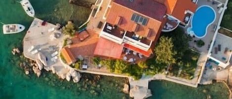 An aerial view of a coastal property with a house, a swimming pool, and several boats docked at a pier.