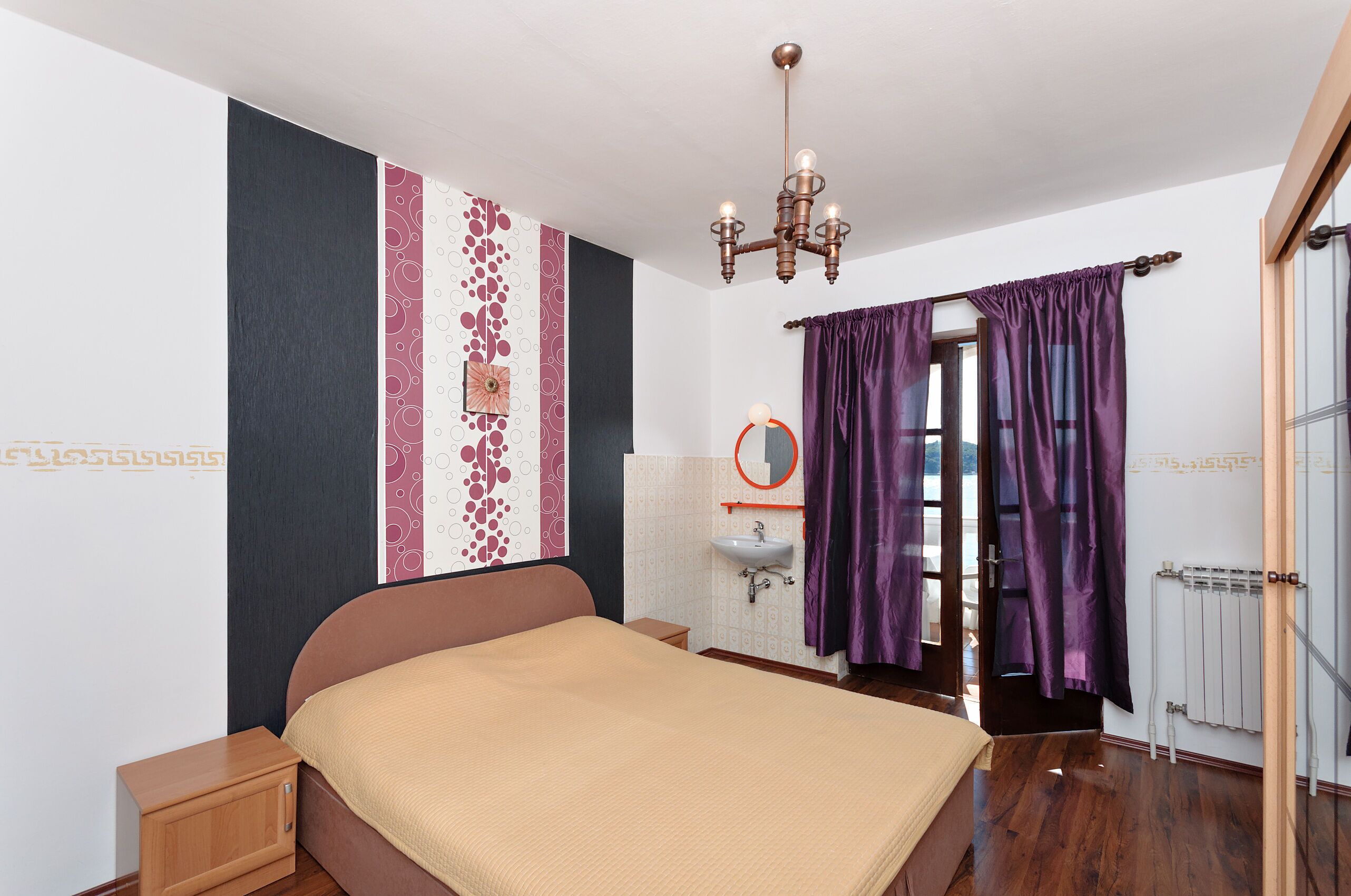 Une chambre à coucher comprenant un lit, une table de chevet, une commode, une fenêtre avec des rideaux, un radiateur et un lustre.