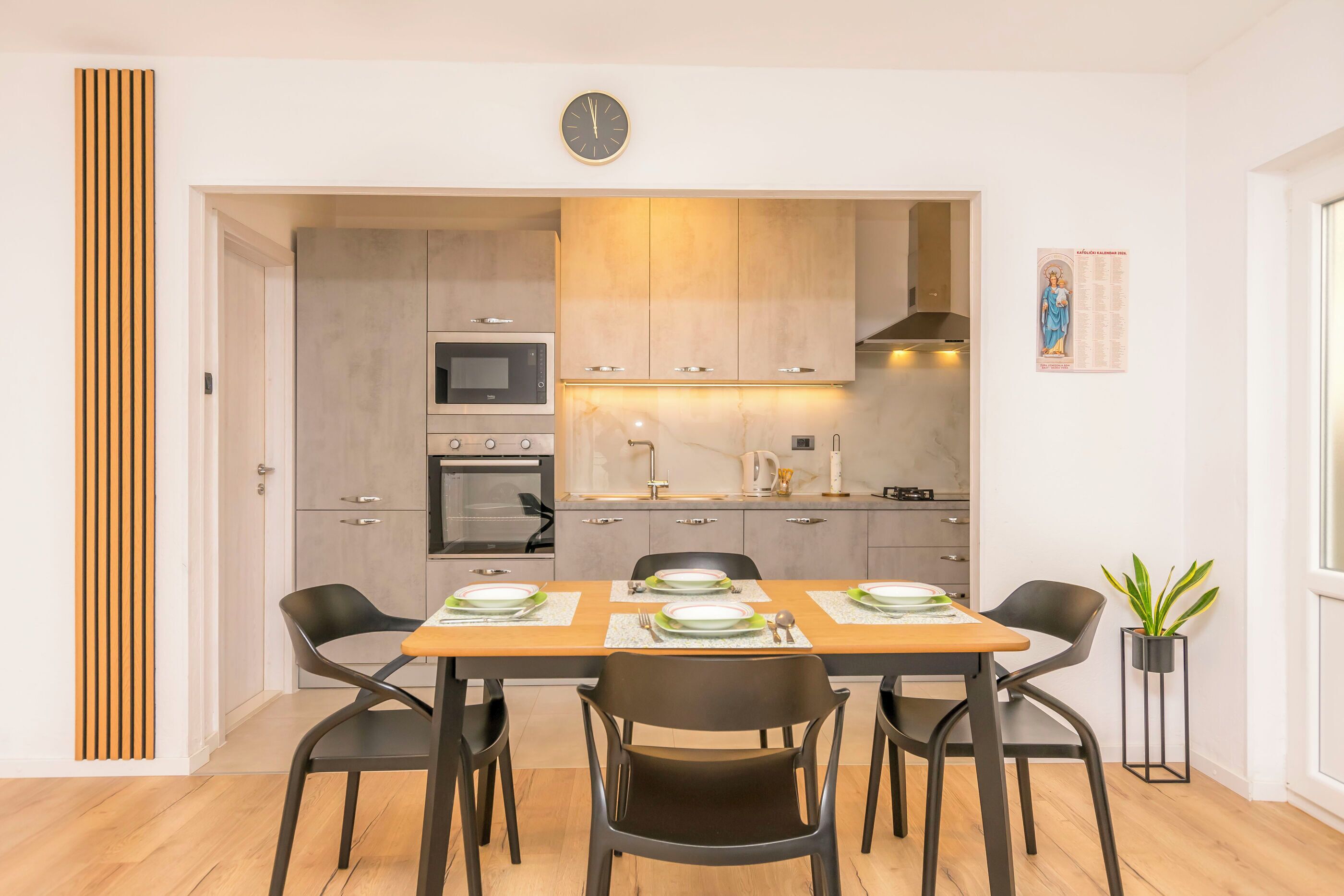 A modern kitchen with a dining table set for four, a clock on the wall, and a potted plant.