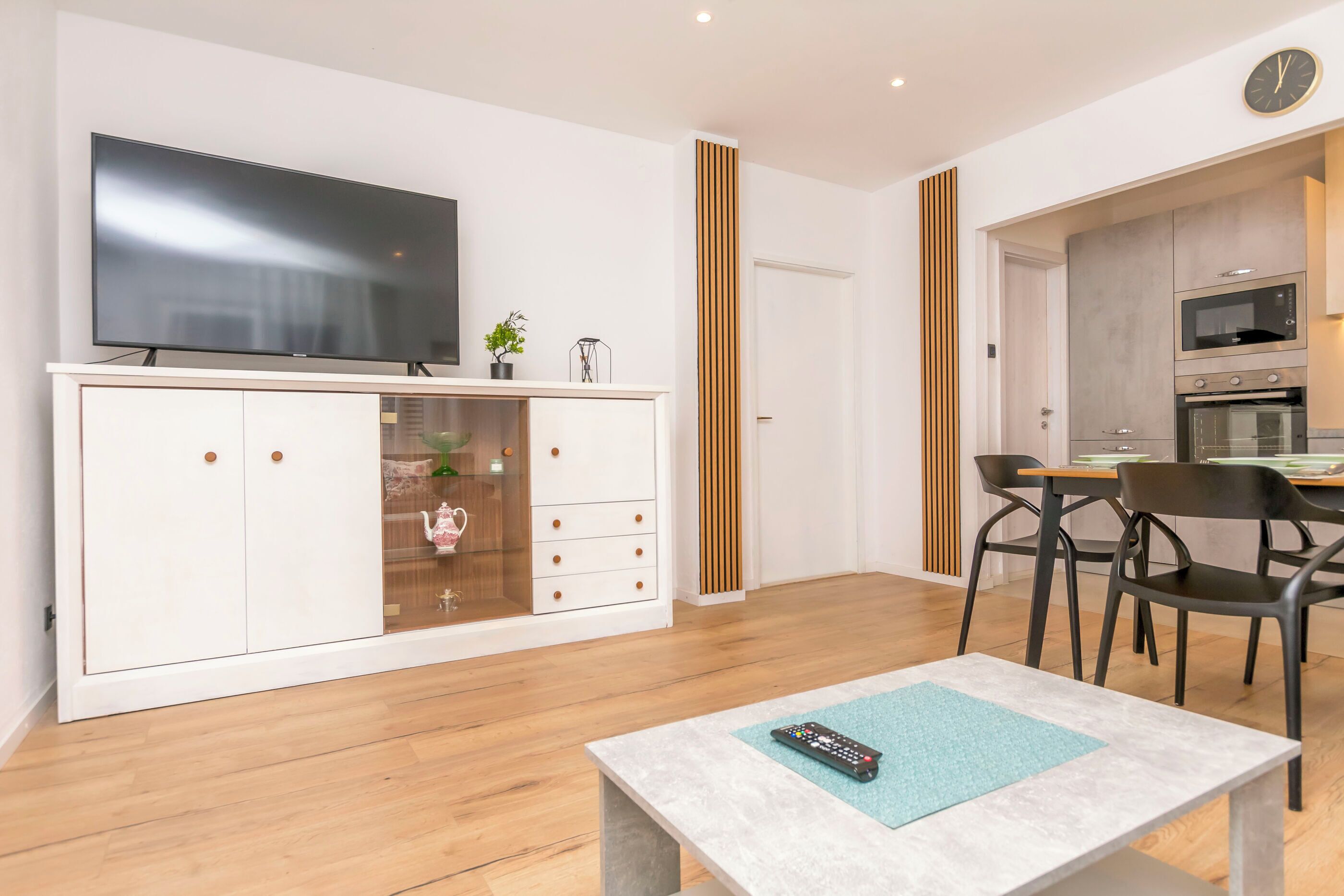 A modern living room with a flat-screen TV, a dining area with a table and chairs, and a kitchen with appliances.