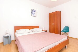 A neatly made bed with a pink blanket, a wooden wardrobe, a small bedside table with a lamp, and a blue chair. - Apartment Mariolina (Baska Voda)