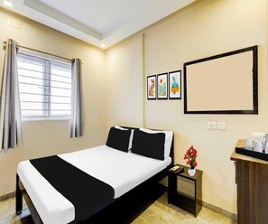 A hotel room with a bed, a nightstand, a desk with a vase of flowers, and a window with curtains. - Classic RK Residency (Coimbatore)