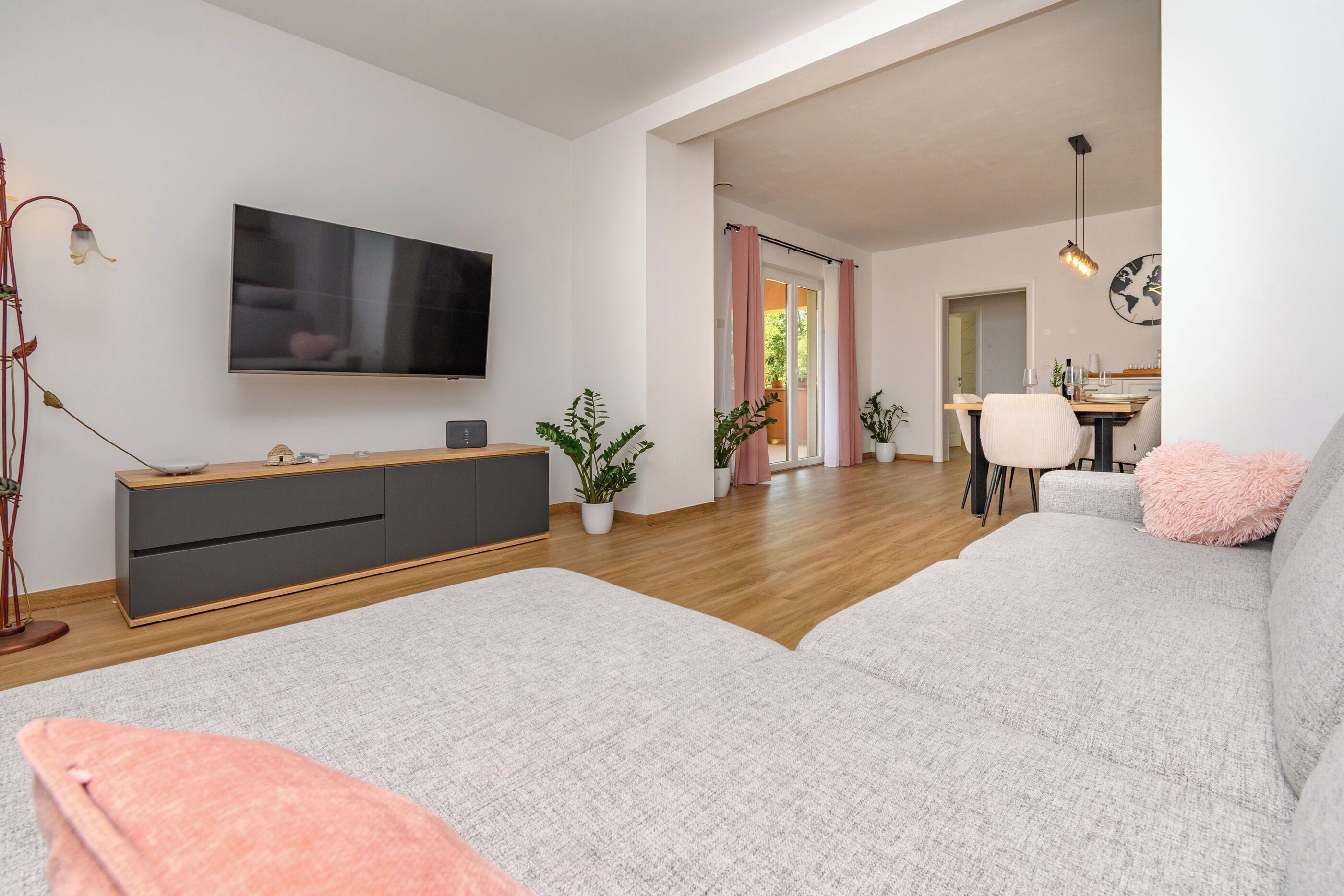 A modern living room with a grey sofa, a flat-screen TV, a wooden cabinet, and a dining area with a table and chairs.