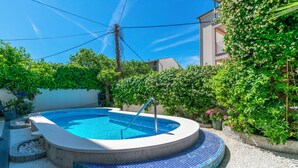 Ein Schwimmbecken mit Fliesenrand, umgeben von Kies und neben einem Gebäude mit weißer Fassade und einem grünen Heckenstreifen.