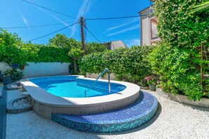 A swimming pool with a tiled edge, surrounded by a pebble ground, and adjacent to a building with a white exterior and a green hedge. - Apartments Wellness&spa (Dugi Rat)