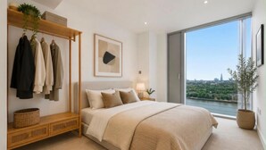 A bedroom with a large bed, a bedside table, a hanging clothes rack, and a window with a city view. - Canary Wharf Skyline Retreat (London)