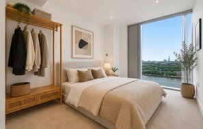 A bedroom with a large bed, a bedside table, a hanging clothes rack, and a window with a city view. - Canary Wharf Skyline Retreat (London)