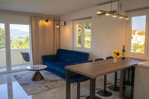 A modern living room with a blue sofa, a round coffee table, and a dining table with chairs. There is a window with a view of greenery and a mountain. - Apartment Blue Velvet Apartment (Lumbarda)