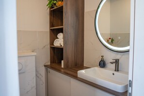 A bathroom with a white sink, a round mirror, a wooden shelf with towels, and a marble wall. - Apartment Blue Velvet Apartment (Lumbarda)