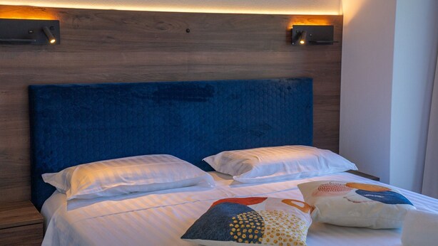 Cama com cabeceira azul, roupa de cama branca e almofadas decorativas.