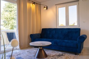 Quarto com sofá azul, mesa redonda, janela com cortinas e vista para árvores.
