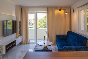 Uma sala de estar moderna com televisão de tela plana, um sofá azul, uma mesa de centro redonda com copos de vinho e uma varanda com cortinas.
